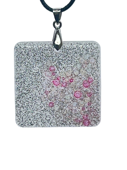 A square-shaped resin pendant densely covered in fine silver glitter, with a cluster of clear and rose-pink bubble-bead inclusions drifting near the center. It hangs from a silver-toned bail on a black braided cord against a pale background.