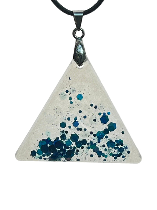 A triangular resin pendant in frosted white, clean at the top point, gathering a dense cluster of sapphire, teal, and midnight blue confetti hexagons toward the base. It hangs from a gunmetal bail on a black braided cord against a pale background.