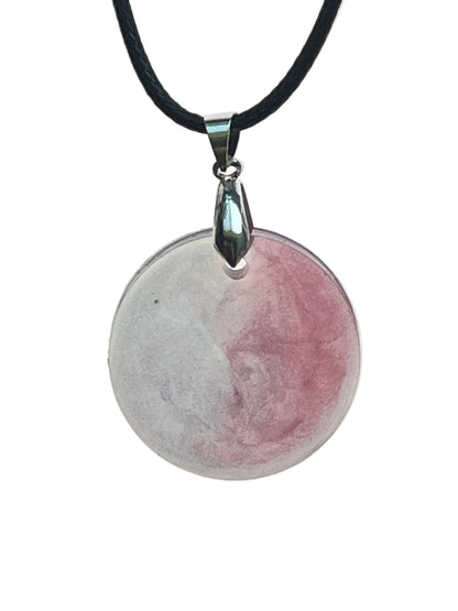 A round disc-shaped resin pendant split between a soft misty grey-white half and a dusty rose-mauve half, meeting in a clean line down the center. It hangs from a gunmetal bail on a black braided cord against a pale background.