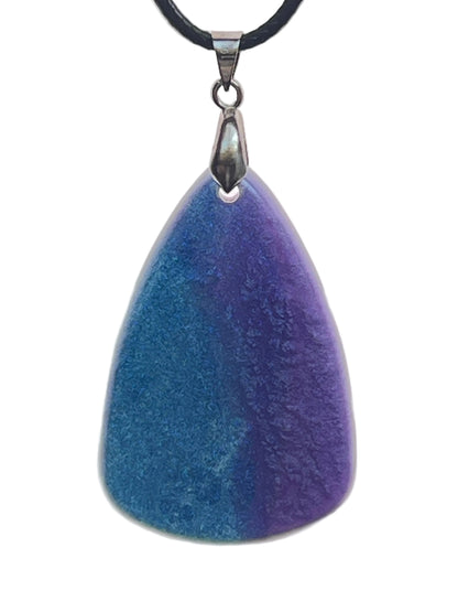 An organic, rounded triangular-shaped resin pendant with a gradient blending teal-blue on one side into deep violet and indigo on the other. It hangs from a silver-toned bail on a black braided cord against a pale background.
