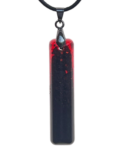 A slender vertical bar-shaped resin pendant with a crimson-red edge at the top that deepens into a near-black void filling the rest of the piece, with fine dark speckles scattered throughout. It hangs from a gunmetal bail on a black braided cord against a pale background.