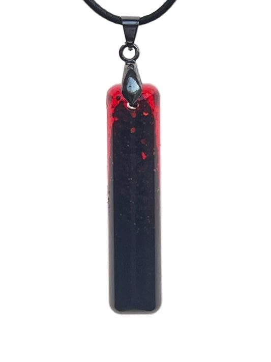 A slender vertical bar-shaped resin pendant with a crimson-red edge at the top that deepens into a near-black void filling the rest of the piece, with fine dark speckles scattered throughout. It hangs from a gunmetal bail on a black braided cord against a pale background.