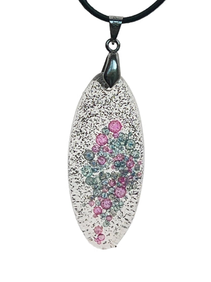 An elongated oval-shaped resin pendant, clear with fine black glitter at the edges, and a cluster of rose-pink and dove-grey bubble-bead inclusions gathered densely at the center. It hangs from a gunmetal bail on a black braided cord against a pale background.