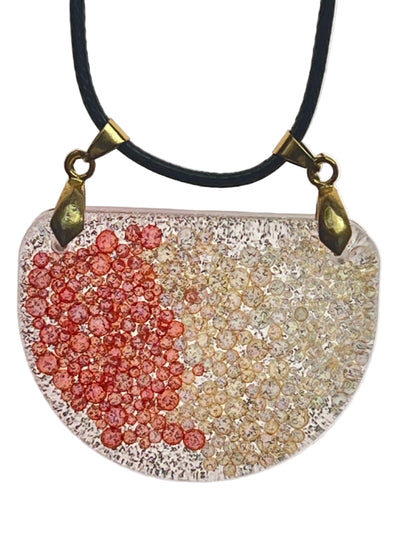 A half-circle (half-moon) shaped resin pendant with clusters of coral-red bubble-beads on one side and champagne-gold bubble-beads on the other, over a clear base with fine dark speckle. It hangs from two antiqued brass bails on a black braided cord against a pale background.