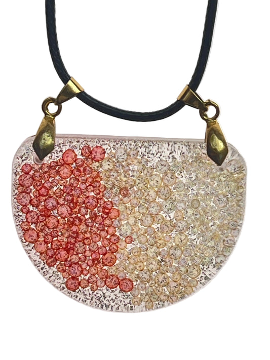 A half-circle (half-moon) shaped resin pendant with clusters of coral-red bubble-beads on one side and champagne-gold bubble-beads on the other, over a clear base with fine dark speckle. It hangs from two antiqued brass bails on a black braided cord against a pale background.