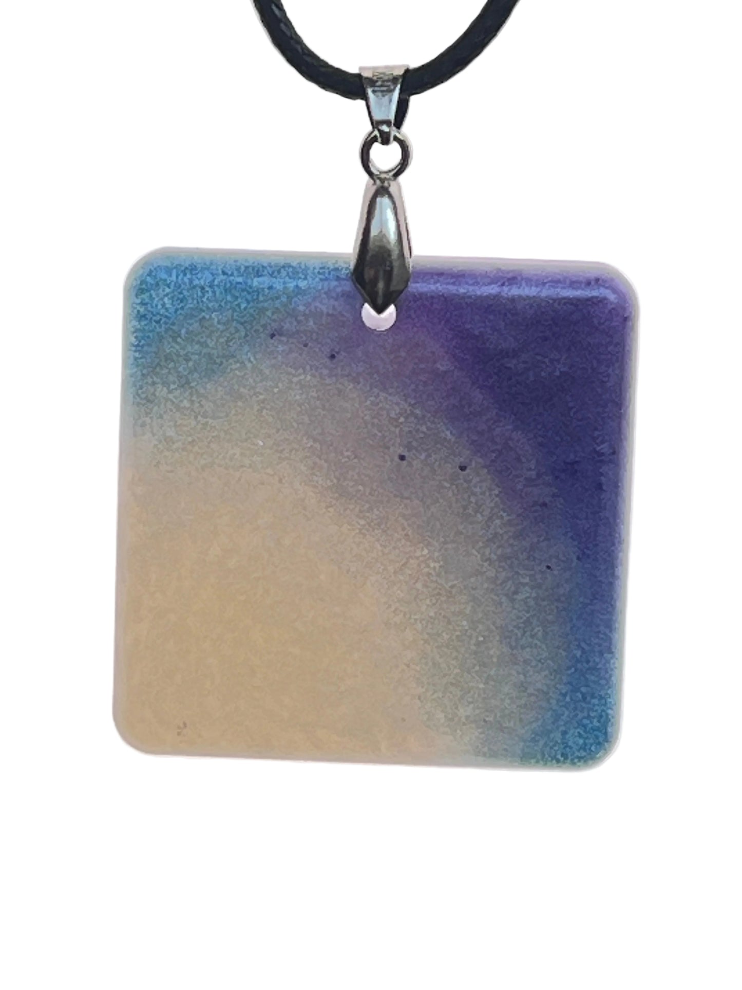 A square resin pendant with a soft gradient from sandy beige in the lower left to sky blue and deep violet-indigo sweeping in from the upper corners. It hangs from a silver-toned bail on a black braided cord against a pale background.