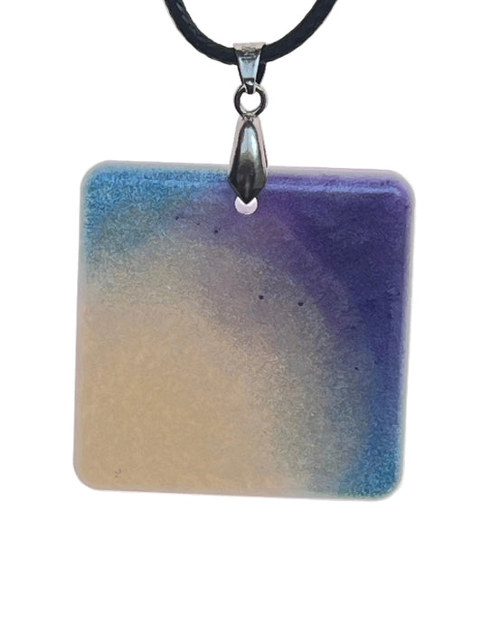 A square resin pendant with a soft gradient from sandy beige in the lower left to sky blue and deep violet-indigo sweeping in from the upper corners. It hangs from a silver-toned bail on a black braided cord against a pale background.