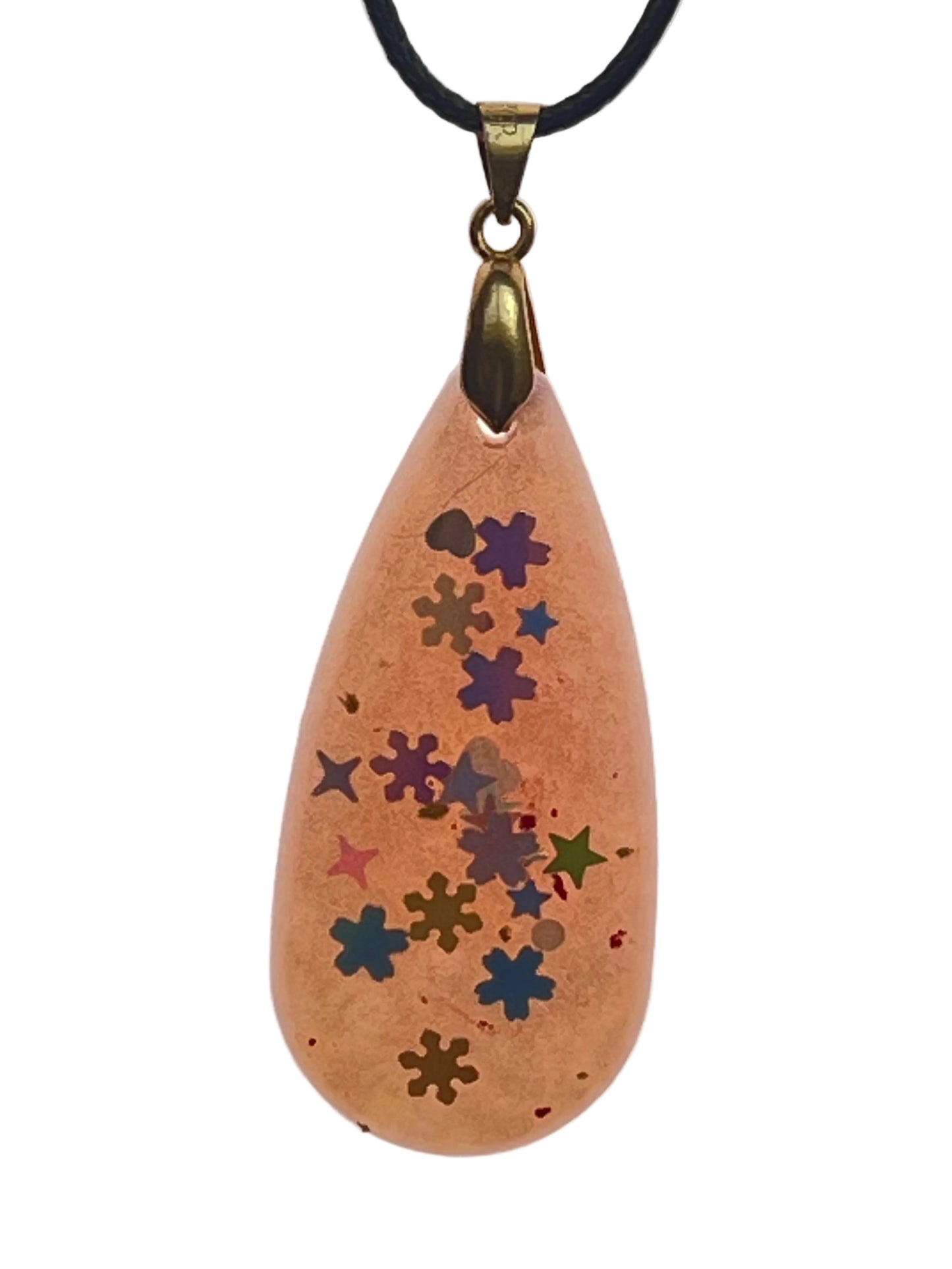 A teardrop-shaped resin pendant in warm peachy-tan with a trail of star and snowflake-shaped confetti in indigo, teal, olive, pink, and blue scattered down the center. It hangs from an antiqued brass bail on a black braided cord against a pale background.