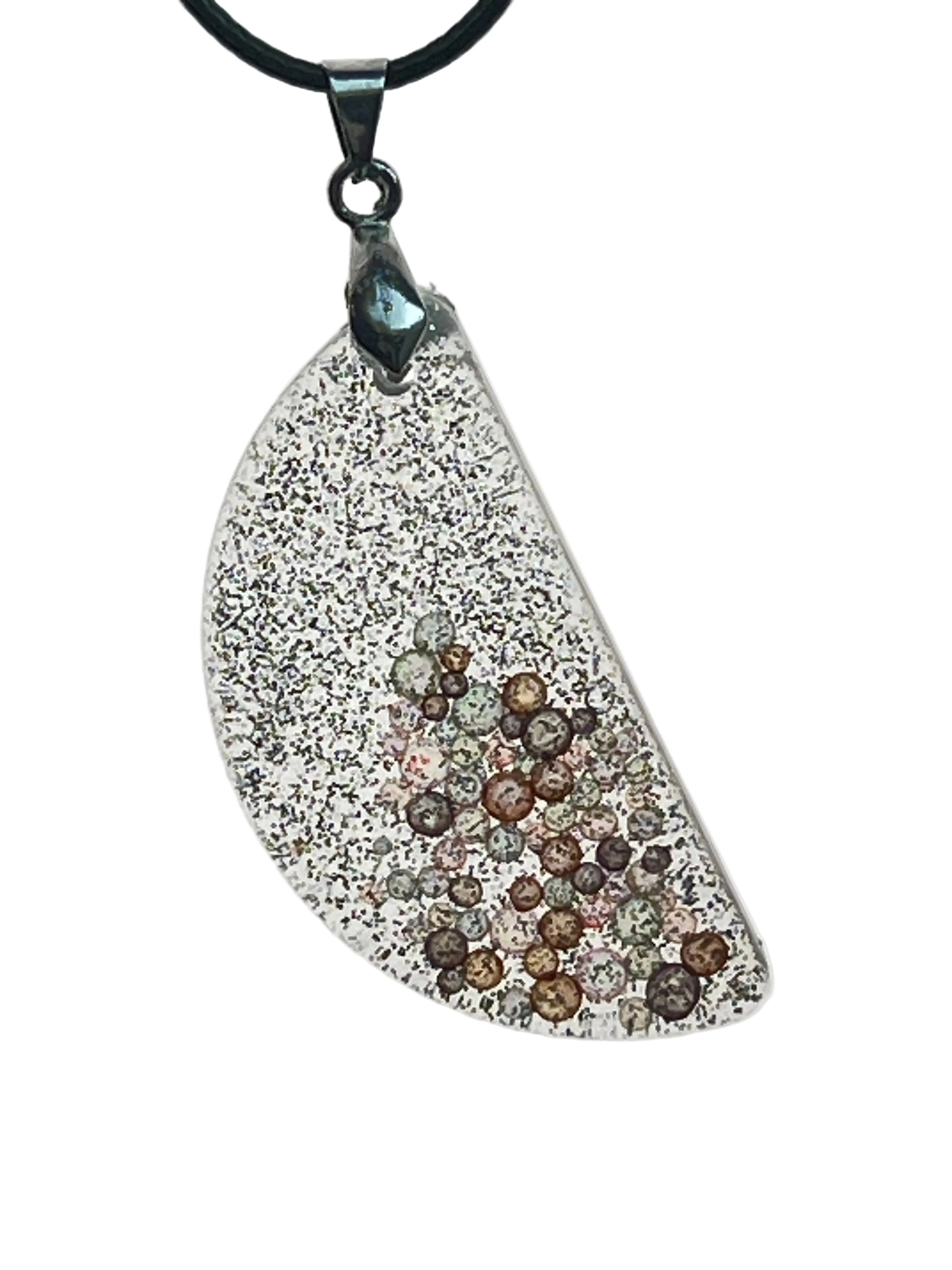 An asymmetrical, wing-shaped resin pendant covered in fine silver glitter, with a cluster of amber, copper, blush, and grey bubble-bead inclusions gathered toward the lower portion. It hangs from a gunmetal bail on a black braided cord against a pale background.