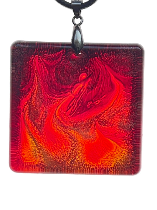 A square resin pendant with a dynamic, flame-like swirling pattern in crimson, burnt orange, and gold, forming a rising winged phoenix-like shape at the center, deepening to near-black at the corners. It hangs from a gunmetal bail on a black braided cord against a pale background.