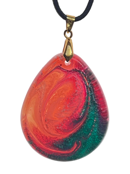A full teardrop-shaped resin pendant with a swirled, eye-like pattern of molten orange and crimson at the center, encircled by rings shifting into deep emerald green toward the base. It hangs from an antiqued brass bail on a black braided cord against a pale background.