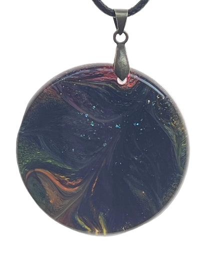 A large round resin pendant with a deep, swirling near-black base dusted with fine glitter, and glimpses of amber, orange, crimson, and jade green along the edges. It hangs from a gunmetal bail on a black braided cord against a pale background.