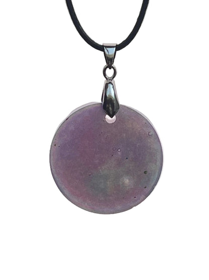 A large round resin pendant in soft violet-grey with subtle sage-green and slate undertones and small bubble-like inclusions. It hangs from a silver-toned bail on a black braided cord against a pale background.