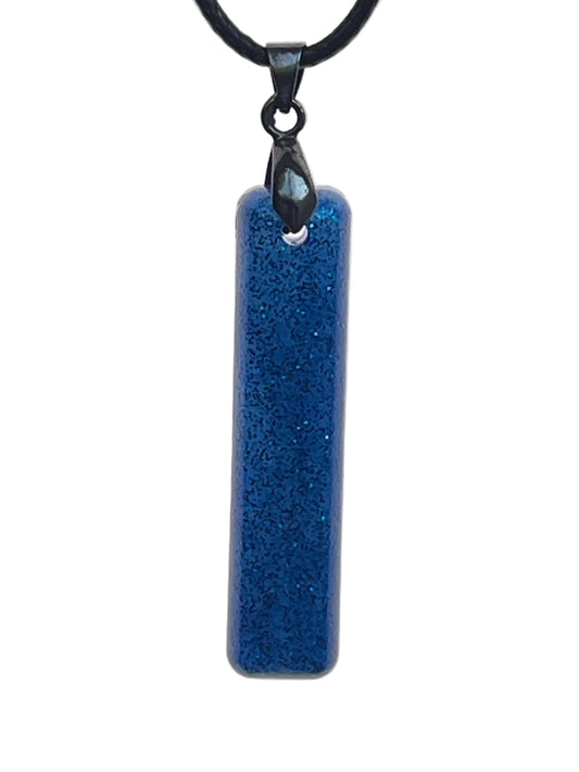 A slender vertical bar-shaped resin pendant in dense, deep navy-sapphire blue with fine sparkling blue and silver glitter throughout. It hangs from a gunmetal bail on a black braided cord against a pale background.
