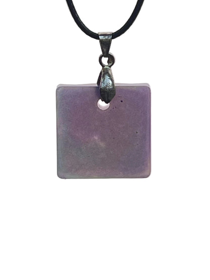 A square resin pendant in soft violet-grey with subtle cloud-like variations of muted plum throughout. It hangs from a gunmetal bail on a black braided cord against a pale background.